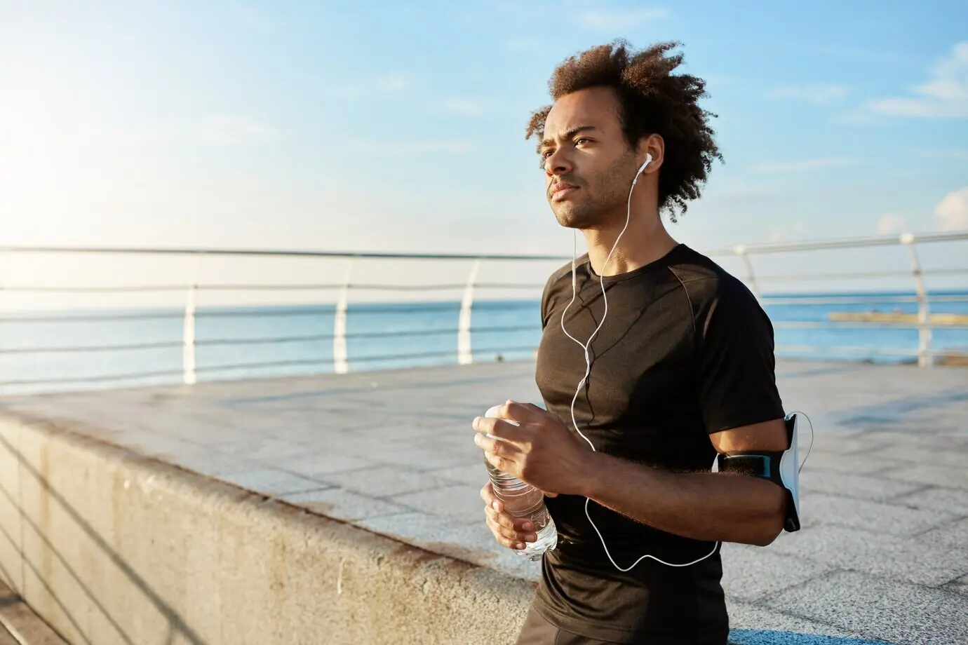 Stylish male jogger with a bushy hairstyle looking straight ahead in the morning, enjoying sports activities. A fit man wearing earphones, holding a bottle of water in his hands, taking a break in the middle of training.